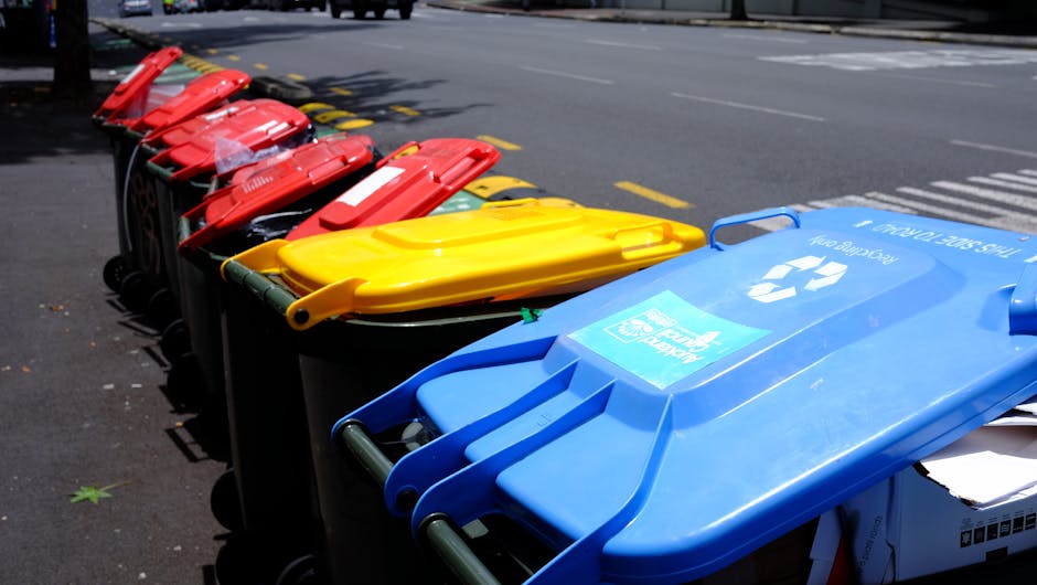 A row of large, rectangular wheelie bins positioned outdoors along a paved pavement, with each bin featuring a matching lid in different colors including green, red, and blue. The green bin in the for